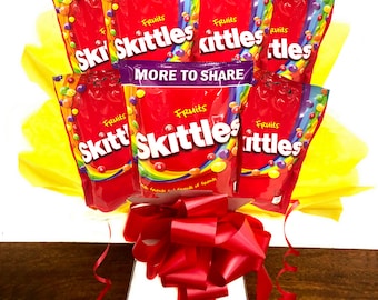 May include: A candy bouquet featuring several red bags of Skittles arranged with yellow tissue paper and a large red bow. The bags have the word "Fruits" and "Skittles" printed on them, with the phrase "MORE TO SHARE" on one bag.