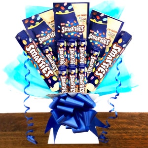 May include: A bouquet of Smarties chocolate bars wrapped in blue and white tissue paper with a blue ribbon. The bouquet is sitting on a wooden surface.