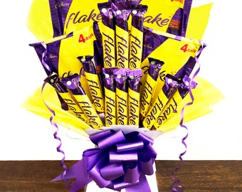 May include: A bouquet of Cadbury Flake chocolate bars wrapped in yellow paper with a purple ribbon. The bouquet is arranged in a white vase. The chocolate bars are arranged in a fan shape and are all facing the same direction. The bouquet is sitting on a wooden table.