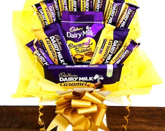 May include: A gift basket filled with Cadbury Dairy Milk Caramel chocolate bars and Caramel Nibbles. The arrangement is wrapped in yellow tissue paper and tied with a gold bow. The chocolate bars are in purple and gold packaging.
