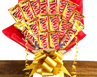 May include: A bouquet of chocolate bars wrapped in gold foil with the brand name "TWIX" printed on each bar. The bouquet is arranged in a white vase with a gold ribbon bow. The bouquet is sitting on a wooden surface.