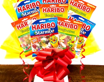 May include: A colorful gift arrangement featuring multiple bags of Haribo gummy candies. The bags are orange and blue, with the Haribo logo and product names like "Starmix" and "Tangfastics". The arrangement is wrapped in yellow tissue paper and tied with a large red bow.