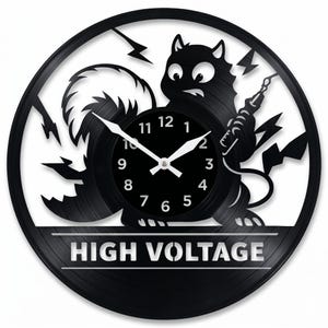 Peut inclure: Horloge en vinyle noir avec un chat de dessin animé tenant un pistolet à tatouer, entouré d'éclairs. Le cadran de l'horloge est noir avec des chiffres et des aiguilles blancs. Le texte "HIGH VOLTAGE" est affiché en bas.