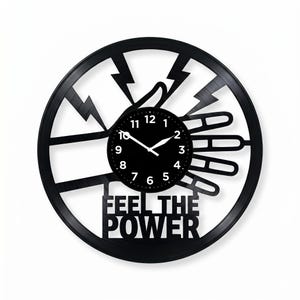 Peut inclure: Horloge murale noire au design unique. L'horloge représente une main avec des éclairs et les mots "FEEL THE POWER". Le cadran de l'horloge est noir avec des chiffres et des aiguilles blancs.