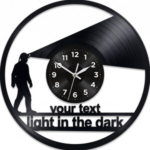 May include: Black vinyl record wall clock with a circular design. The clock features a silhouetted figure walking with a light, a clock face, and the text "your text light in the dark". The clock hands are white.