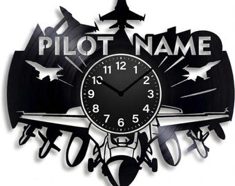 Könnte beinhalten: Schwarze Wanduhr mit Pilotenmotiv, aus einer Schallplatte geschnitten. Die Uhr hat ein zentrales Zifferblatt mit weißem Rand und schwarzen Ziffern, umgeben von Silhouetten von Kampfflugzeugen und den Worten "PILOT NAME".