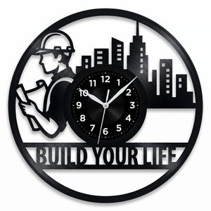 May include: Black wall clock with a circular design featuring a silhouette of a construction worker and a cityscape. The clock face is black with white numerals and hands. The words "BUILD YOUR LIFE" are at the bottom.