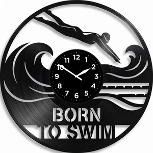 Könnte beinhalten: Eine runde Wanduhr aus einer schwarzen Schallplatte mit einer ausgeschnittenen Silhouette eines Schwimmers, der in Wellen eintaucht. Das Zifferblatt hat weiße Zahlen und Zeiger. Der Text BORN TO SWIM ist am unteren Rand der Uhr ausgeschnitten.