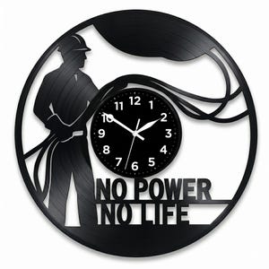 May include: Black wall clock with a silhouette of an electrician holding cables, with the text "NO POWER NO LIFE". The clock face is black with white numbers and hands. The clock is round.