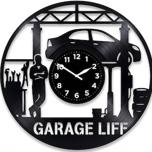 Puede incluir: Reloj de pared negro con temática de garaje. El reloj presenta un coche en un elevador, un mecánico y las palabras "GARAGE LIFE". La esfera del reloj es negra con manecillas y números blancos.