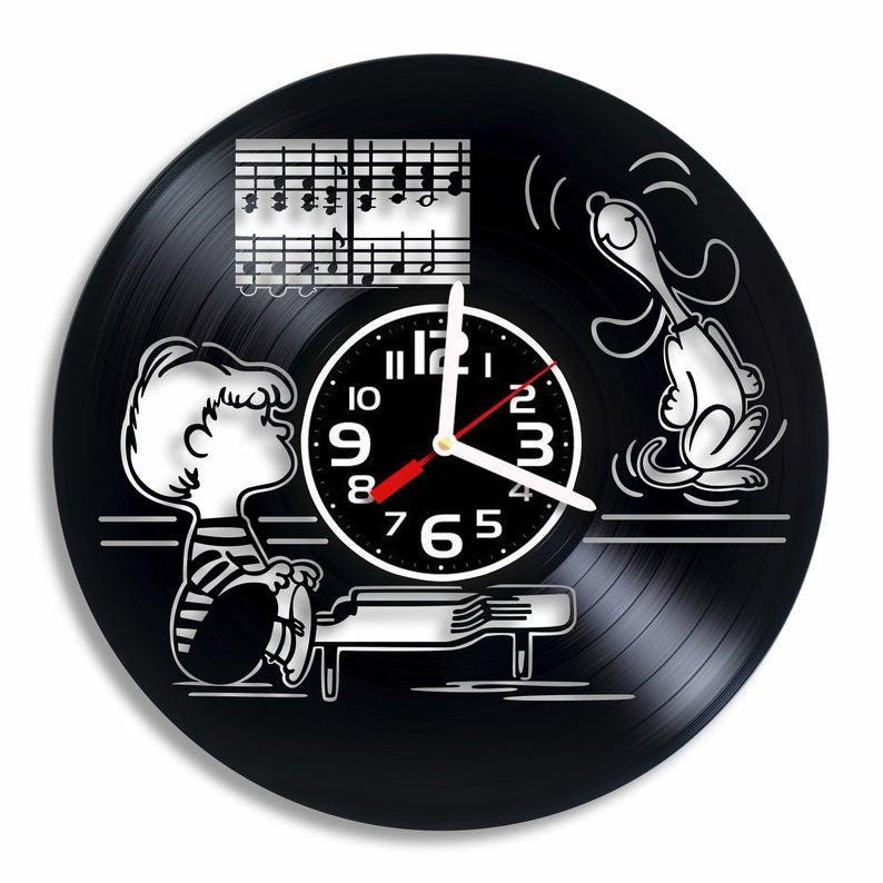 May include: Black vinyl record clock featuring Peanuts characters Charlie Brown and Snoopy. Charlie Brown is playing piano with musical notes above him; Snoopy is singing.  The clock face is centrally located with black and white numbers.  The overall design is minimalist and stylish, suitable for music lovers and Peanuts fans.