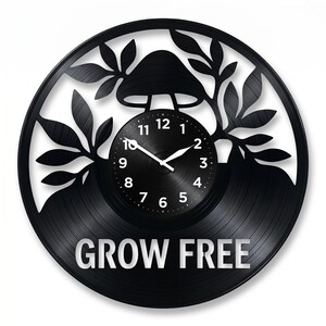 Puede incluir: Reloj de vinilo negro con diseño de hongo y hojas. La esfera del reloj es negra con números y manecillas blancas. Las palabras "GROW FREE" están impresas en blanco en la parte inferior del reloj.