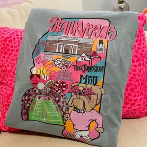 May include: A grey t-shirt featuring a colorful graphic print of the state of Mississippi. The design includes a building, a bulldog wearing star-shaped glasses, a football field, sports equipment, and text reading Stankvegas, Hail State, and MSU.