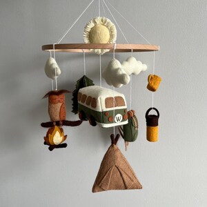 May include: A felt mobile with a camping theme. It features a bus, owl, campfire, tent, clouds, sun, backpack, and a mug. The mobile hangs from a wooden ring with white strings, making a charming nursery decoration.