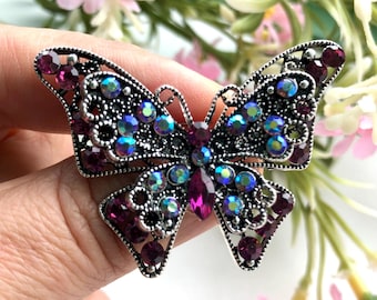 May include: A butterfly-shaped brooch featuring a dark silver-toned metal frame with intricate cut-out details. The wings are embellished with a mix of deep purple round rhinestones and iridescent, multi-coloured crystals that shimmer with blue and green hues.