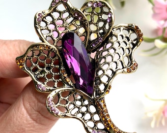 May include: A flower-shaped brooch with an antique gold-tone metal filigree frame. The centre features a large, elongated, faceted purple crystal. The petals are accented with small, round, sparkling amber and light pink crystals along the edges.