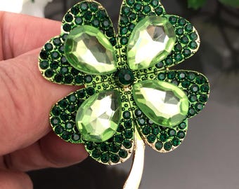 May include: A four-leaf clover brooch featuring a gold-tone metal frame, four large pear-shaped light green crystals at the centre of each leaf, and numerous small dark green rhinestones covering the surface of the clover leaves.