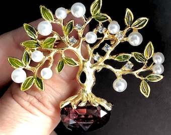 May include: Gold-coloured tree brooch featuring green enamel leaves, faux pearls, and clear crystals. The tree trunk is black, and the base is a faceted, deep red gemstone. A decorative jewellery item.