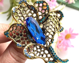 May include: A gold-tone metal brooch shaped like a flower with an openwork filigree design. The centre features a large, elongated, faceted dark blue crystal, while the petals are accented with numerous small, round, light blue rhinestones.