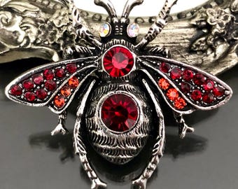 May include: A silver-toned insect brooch featuring a textured metal body, two iridescent eyes, and large red circular crystals set in the thorax and abdomen. The wings are embellished with numerous small red and orange crystals in a gradient pattern.