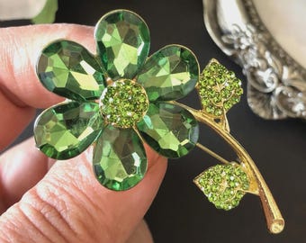 May include: A gold-coloured flower brooch featuring large, faceted green crystal petals and a smaller, round, green crystal centre. The brooch includes a stem with green crystal leaves.