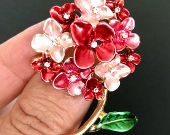 May include: A floral brooch with red and pink flowers, each with a rhinestone. The brooch has a gold-tone stem and a green leaf. The flowers are arranged in a bouquet shape, creating a vibrant accessory. The brooch is a decorative item.