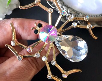 May include: A gold-toned spider brooch featuring a large, clear, pear-shaped crystal for the abdomen and a round, iridescent, multi-coloured crystal for the thorax. The eight legs are accented with small, clear, and iridescent round crystals.