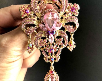 May include: An elaborate gold-coloured brooch adorned with pink and purple gemstones. The brooch has a large, teardrop-shaped pink stone in the centre, encircled by smaller pink and purple crystals. Dangling iridescent stones are suspended from the base.