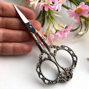 May include: Ornate, vintage-style scissors with a silver blade and decorative bronze handles. The handles feature floral and scrollwork designs. The scissors are designed for fine detail work, such as embroidery or crafting.