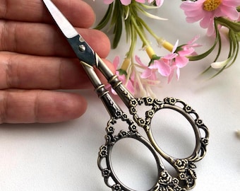 May include: Vintage-style scissors with a silver blade and decorative bronze handles. The handles feature floral and scrollwork designs. These ornate scissors are designed for fine detail work, such as embroidery or crafting.
