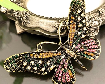 May include: A butterfly-shaped brooch featuring a gold-tone metal frame with an intricate cutout design. The wings are embellished with small, multi-coloured rhinestones in shades of green, pink, orange, and white, arranged in a patterned layout.