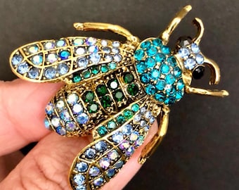 May include: A gold-coloured bee brooch, embellished with shimmering blue, green, and clear crystals. The insect-shaped pin features intricate details, including antennae and wings, creating a glamorous accessory.