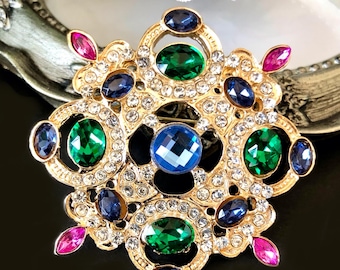 May include: A gold brooch with an elaborate design, set with blue, green, and pink gemstones. The brooch is further adorned with numerous small, clear crystals. The central focus is a large blue gem. The design is symmetrical and would suit formal wear.