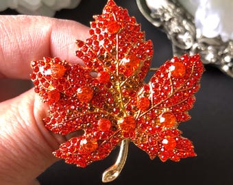 May include: A maple leaf brooch featuring a gold-tone metal base covered in vibrant red and orange rhinestones. The leaf design includes several larger, round, faceted orange crystals interspersed among the smaller red stones, with a smooth gold stem.