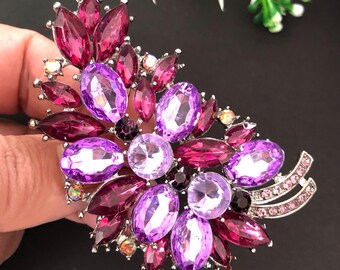 May include: A silver brooch with a floral design. The brooch features a cluster of purple and magenta gemstones in various shapes. The gemstones are set in a silver metal base, creating a sparkling and elegant accessory for a jacket or coat.