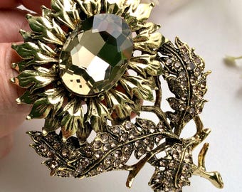 May include: A gold-toned metal brooch shaped like a sunflower with a large, oval, faceted champagne-coloured central stone. The petals are textured, and the stem and leaves are embellished with small, clear, sparkling rhinestones throughout.