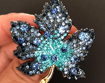 May include: A maple leaf brooch featuring a gradient of blue, teal, and turquoise crystals. The leaf has a dark metal frame with a smooth gold-toned stem at the base. The surface is densely covered in faceted round crystals of varying sizes.