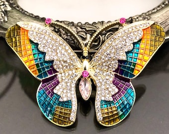 May include: A butterfly brooch featuring gold-tone metal, clear crystal pavé accents, and vibrant enamel sections in shades of orange, teal, and purple. The design includes a central pink teardrop crystal and two small pink crystal antennae tips.
