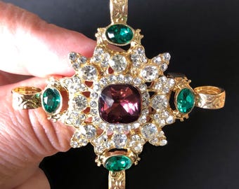 May include: A gold-tone metal cross pendant featuring a large central square purple gemstone, four oval green gemstones at the ends of the arms, and numerous small clear rhinestones set throughout the ornate, textured metal frame.