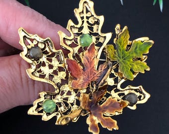 May include: A gold-tone metal brooch shaped like a cluster of maple leaves. The design features openwork filigree detailing, two small green cabochon stones, two small grey cabochon stones, and two smaller enamel-coated leaves in orange and green.