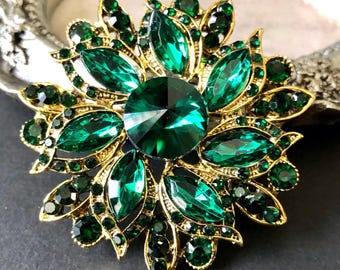 May include: A gold-coloured brooch with a floral design, featuring emerald green crystals. The brooch has a large, faceted centre stone surrounded by smaller, leaf-shaped crystals and additional smaller stones.