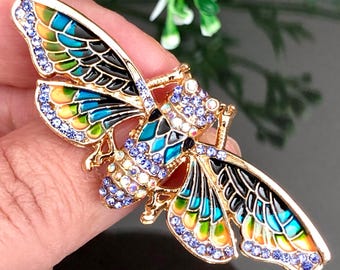May include: A gold insect brooch with detailed design. The wings are decorated with blue, green, yellow, and black enamel, and are embellished with small, sparkling blue crystals. The brooch is designed with a detailed insect body.