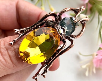 May include: A bronze insect brooch featuring a large, faceted yellow gemstone body and a smaller, faceted blue-green gemstone head. The brooch has detailed legs and antennae. The background is soft and blurred.