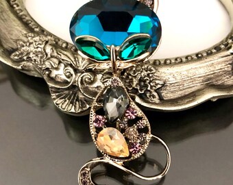 May include: A cat-shaped brooch crafted from gold-tone metal, featuring a large oval teal crystal head with pointed ears. The body is an openwork design adorned with two teardrop-shaped crystals in grey and peach, accented by small purple rhinestones.