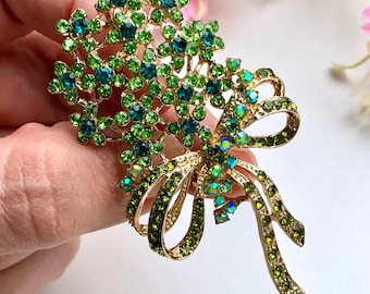 May include: A gold-tone metal brooch shaped like a bouquet of flowers tied with a ribbon. The piece is encrusted with numerous small, sparkling rhinestones in various shades of bright lime green, emerald green, and deep teal blue throughout.