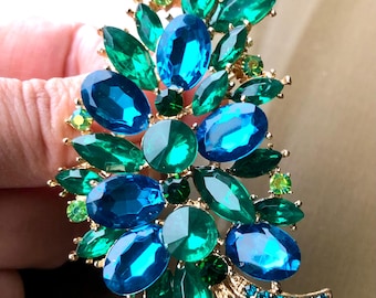 May include: A gold-tone brooch featuring a cluster of faceted oval and round crystals in shades of deep emerald green, bright teal, and royal blue. The arrangement includes smaller lime green accent stones and two curved stems set with tiny blue crystals.
