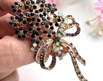 May include: A gold-coloured brooch in the shape of a bouquet with a bow. The brooch features clusters of dark blue and clear crystals, with a bow adorned with purple and light blue crystals. The brooch is held in a hand.