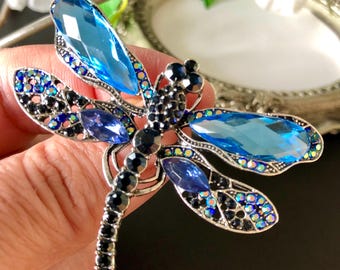 May include: A silver dragonfly brooch with blue and black crystal detailing. The wings have large, light blue crystals and smaller, iridescent stones. The body is detailed with black crystals.
