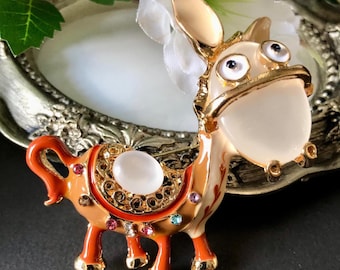 May include: A gold-tone metal brooch shaped like a donkey, featuring a smooth cream-coloured face, orange enamel body, and a saddle decorated with a central white cabochon stone and small multicoloured rhinestones along the edges and legs.