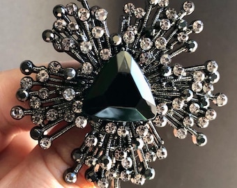 May include: A dark grey brooch with a central triangular, dark green gemstone. The brooch features a radiating design with small, clear and dark grey crystals, creating a sparkling, floral-like pattern. The brooch is a fashion accessory.